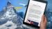 Hand, die Tablet hält, auf dem der FineSolutoins-Newsletter Zoll ohne Grenzen geöffnet ist mit Bergen und einem Flugzeug im Hintergrund