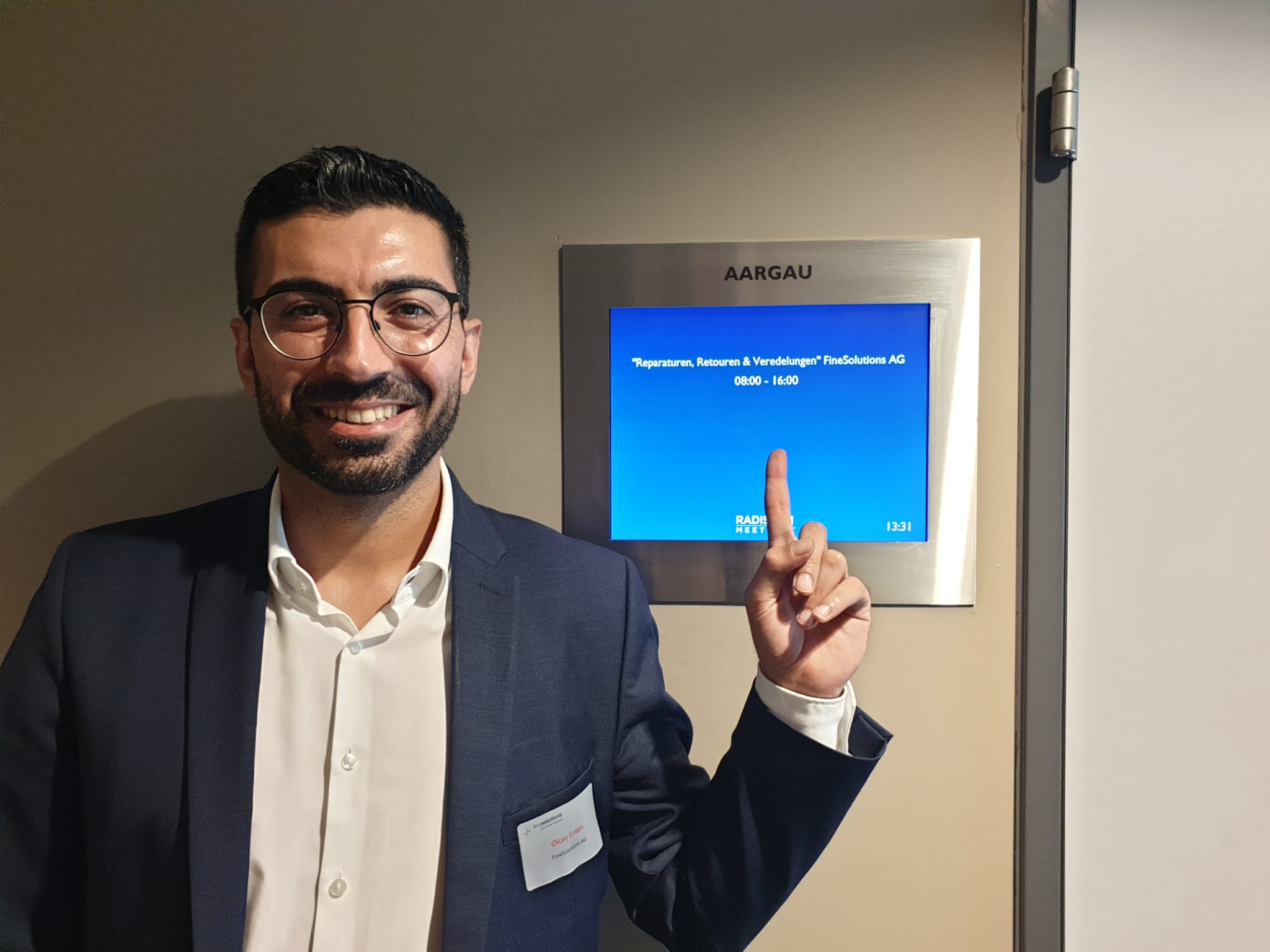 Olcay Erden von FineSolutions steht als Zoll Seminarleiter vor der Anzeigetafel für den Raum Aargau im Radisson Blu und zeigt darauf